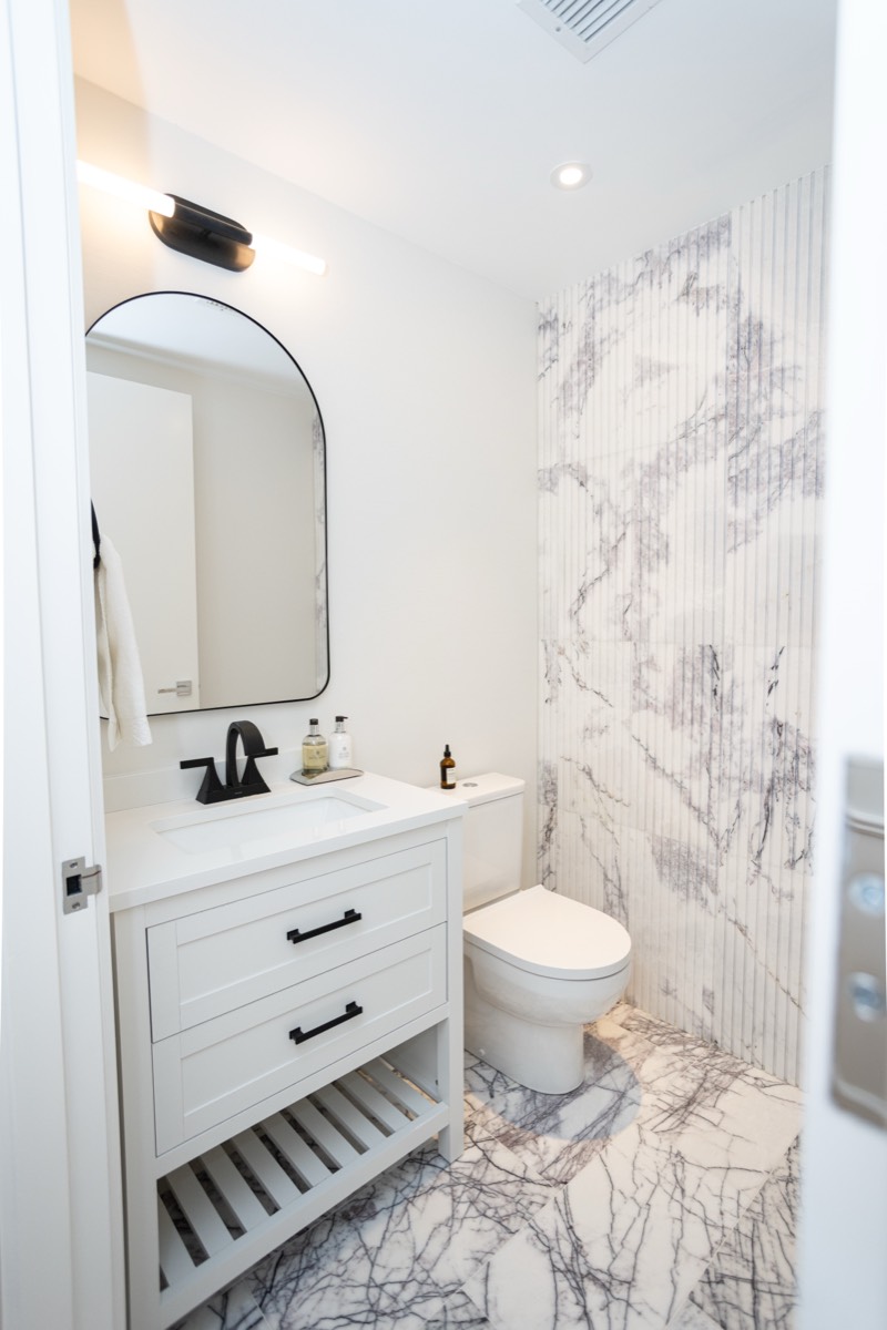 Powder room with a marble feature wall, arched mirror and white vanity with open shelf